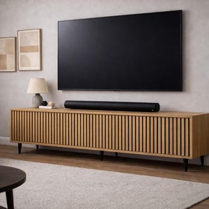 May include: A modern living room setup featuring a large black television mounted above a light wood cabinet with vertical slat detailing. A black soundbar sits on the cabinet. A lamp, artwork, and a potted plant complete the scene.