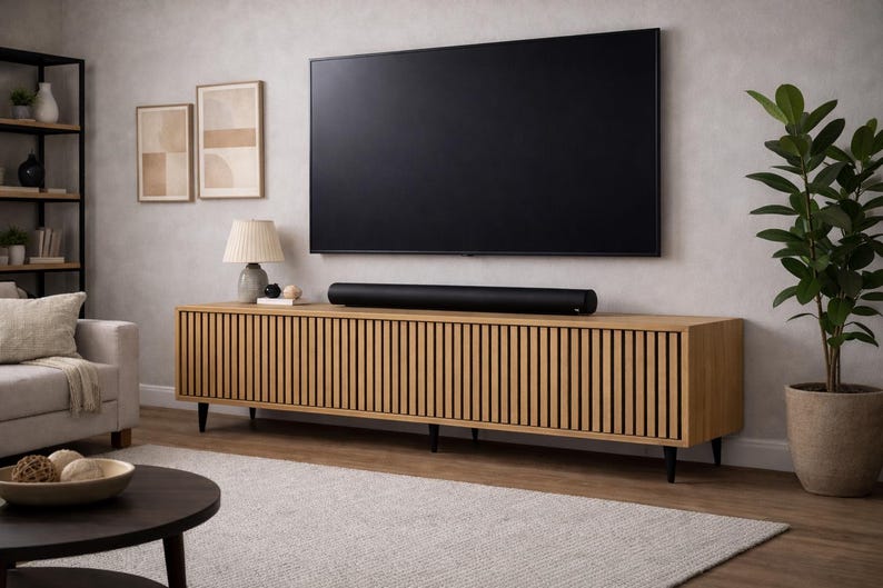 May include: A modern living room setup featuring a large black television mounted above a light wood cabinet with vertical slat detailing. A black soundbar sits on the cabinet. A lamp, artwork, and a potted plant complete the scene.