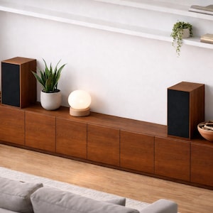 Puede incluir: Una moderna configuraci&oacute;n de sala de estar con un mueble largo de madera oscura con m&uacute;ltiples puertas. Dos altavoces rectangulares con frentes negros se asientan sobre el mueble. Una peque&ntilde;a l&aacute;mpara redonda y plantas en macetas a&ntilde;aden decoraci&oacute;n. Una alfombra y un sof&aacute; de color claro est&aacute;n en primer plano.
