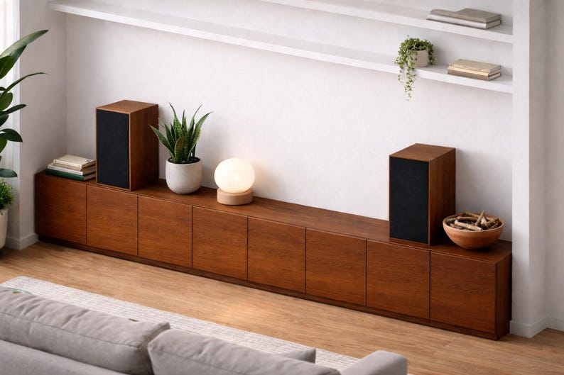 Puede incluir: Una moderna configuraci&oacute;n de sala de estar con un mueble largo de madera oscura con m&uacute;ltiples puertas. Dos altavoces rectangulares con frentes negros se asientan sobre el mueble. Una peque&ntilde;a l&aacute;mpara redonda y plantas en macetas a&ntilde;aden decoraci&oacute;n. Una alfombra y un sof&aacute; de color claro est&aacute;n en primer plano.