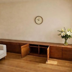 Puede incluir: Un mueble largo de madera con m&uacute;ltiples puertas y estanter&iacute;as abiertas. El mueble es de color marr&oacute;n c&aacute;lido y se encuentra contra una pared de color claro. Un reloj redondo cuelga de la pared encima del mueble. Un jarr&oacute;n de flores blancas est&aacute; encima.
