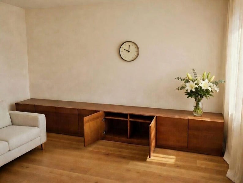 Puede incluir: Un mueble largo de madera con m&uacute;ltiples puertas y estanter&iacute;as abiertas. El mueble es de color marr&oacute;n c&aacute;lido y se encuentra contra una pared de color claro. Un reloj redondo cuelga de la pared encima del mueble. Un jarr&oacute;n de flores blancas est&aacute; encima.