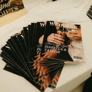 以下が含まれることがあります： 「WEDDING」の文字が白で書かれた黒いウェディング雑誌の束。表紙にはシャンパングラスを持つ2人の写真が写っています。「Best Day Ever ISABELLA & TAYLOR」の文字も確認できます。