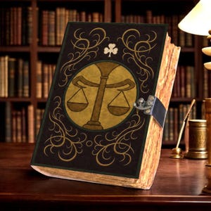 Op de afbeelding: Een zwart hardcover boek met een goudkleurig cirkelvormig embleem van een weegschaal op de voorkant, versierd met gouden krulmotieven en een klein klaverblad-symbool. Het boek is aan de rechterkant afgesloten met een donkere metalen sluiting.