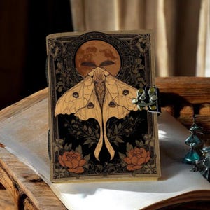 Puede incluir: Un diario de estilo vintage con un dise&ntilde;o de polilla y luna. La portada presenta una ilustraci&oacute;n detallada de una polilla con una luna llena en el fondo, rodeada de elementos florales. El diario est&aacute; asegurado con un cierre met&aacute;lico.