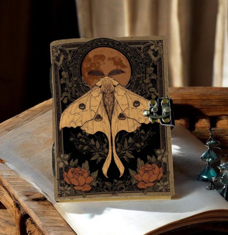 Puede incluir: Un diario de estilo vintage con un dise&ntilde;o de polilla y luna. La portada presenta una ilustraci&oacute;n detallada de una polilla con una luna llena en el fondo, rodeada de elementos florales. El diario est&aacute; asegurado con un cierre met&aacute;lico.
