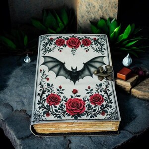May include: A decorative journal with a bat and rose design. The cover features a black bat with outstretched wings, surrounded by red roses and green foliage on a cream background. The journal has a metal clasp and aged-looking pages.