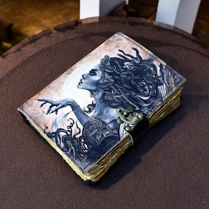 May include: A leather journal with an illustration of a woman with long hair and a dramatic pose. The cover art is in shades of blue and cream, with a brass clasp and a black leather strap. The pages have a vintage, aged appearance, giving it a unique look.