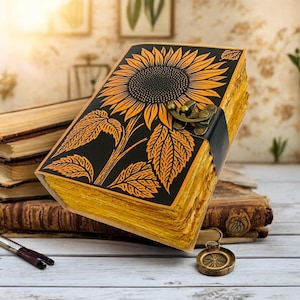 May include: A rectangular journal with a black cover featuring a large yellow sunflower illustration. The journal has thick, textured yellow pages and is secured with a dark metal latch. A small, round, metallic charm hangs from the bottom edge.