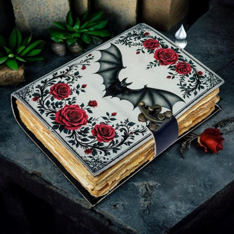 May include: A decorative journal with a bat and rose design. The cover features a black bat with outstretched wings, surrounded by red roses and green leaves on a white background. The journal has a dark blue leather strap and a metal clasp.
