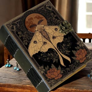 Puede incluir: Un diario decorativo con un dise&ntilde;o de polilla y luna. La portada presenta una gran ilustraci&oacute;n de polilla en tonos beige y marr&oacute;n, sobre un fondo negro con detalles florales. El diario tiene una lomo verde oscuro y un cierre met&aacute;lico.