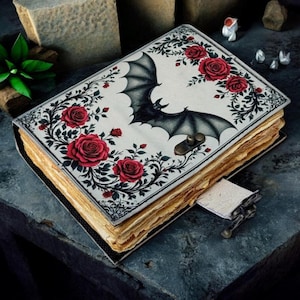 May include: A decorative journal with a bat and rose design. The cover features a black bat with outstretched wings, surrounded by red roses and green vines on a white background. The journal has aged-looking pages and a metal clasp.