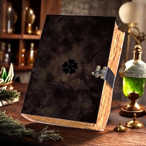 May include: A dark brown, textured journal with a black five-leaf clover emblem centred on the front cover. The book features a metallic, antique-style latch closure and thick, rough-edged pages that appear aged and light tan in colour.