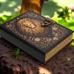 Op de afbeelding: Een zwart dagboek met een gouden metalen sluiting. De voorkant bevat een gedetailleerde illustratie van een leeuwenkop omringd door ingewikkelde bloemmotieven en een kleine maansikkel. De pagina's hebben een verweerd, verouderd uiterlijk.