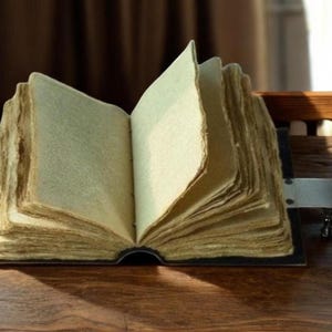 Puede incluir: Un libro abierto de estilo antiguo, con p&aacute;ginas amarillentas. El libro tiene una cubierta oscura y un cierre met&aacute;lico. Las p&aacute;ginas son gruesas y tienen un borde &aacute;spero e irregular, lo que sugiere una calidad vintage. El libro est&aacute; sobre una superficie de madera.