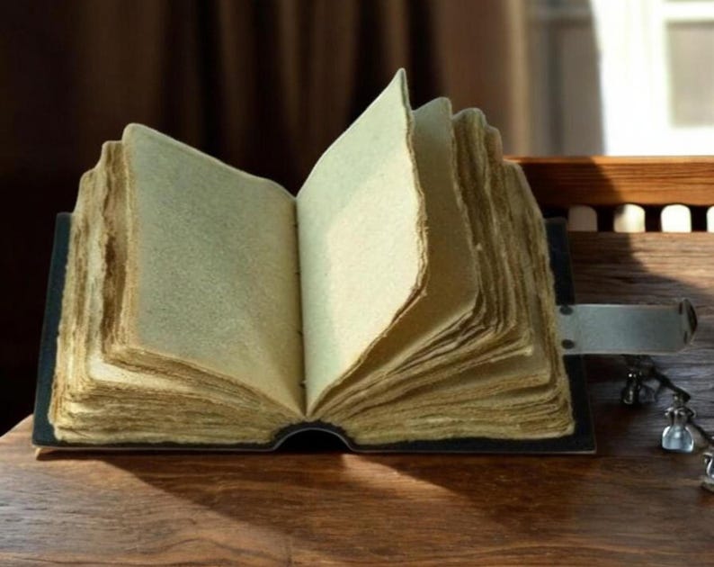 Puede incluir: Un libro abierto de estilo antiguo, con p&aacute;ginas amarillentas. El libro tiene una cubierta oscura y un cierre met&aacute;lico. Las p&aacute;ginas son gruesas y tienen un borde &aacute;spero e irregular, lo que sugiere una calidad vintage. El libro est&aacute; sobre una superficie de madera.