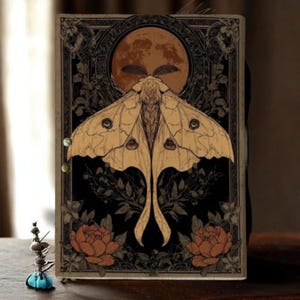 Puede incluir: Un diario decorativo con un dise&ntilde;o de polilla y luna. La portada presenta una gran polilla con alas detalladas, sobre un fondo negro con detalles florales y una luna llena. La paleta de colores incluye tonos beige, negro y naranja.