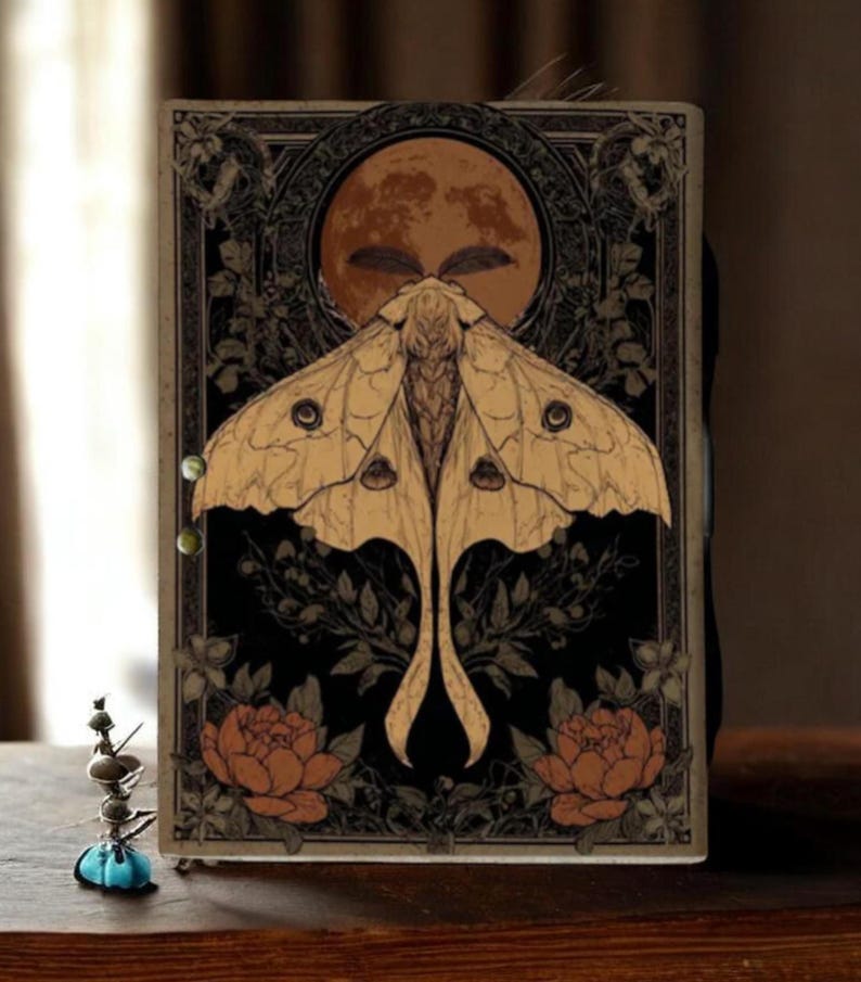 Puede incluir: Un diario decorativo con un dise&ntilde;o de polilla y luna. La portada presenta una gran polilla con alas detalladas, sobre un fondo negro con detalles florales y una luna llena. La paleta de colores incluye tonos beige, negro y naranja.
