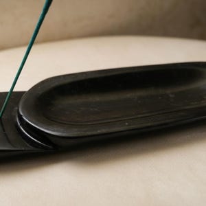Può includere: Un porta incenso nero di forma ovale con una base abbinata. Un bastoncino di incenso verde è inserito nel supporto. Il supporto è realizzato in un materiale liscio e scuro e poggia su una superficie di colore chiaro.