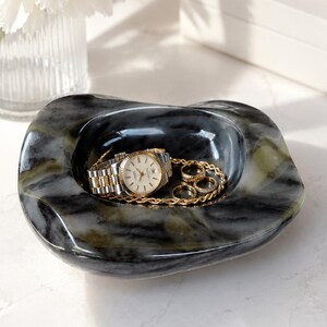 May include: A polished, irregular-shaped dark gray and black marble catch-all bowl with visible veining. Inside the bowl rests a two-tone silver and gold metal wristwatch with a round face, a gold chain necklace, and three gold rings of varying sizes.