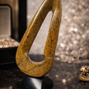 Pode incluir: Uma escultura abstrata em forma de gota, feita de pedra castanho-dourada salpicada. É montada numa base preta geométrica. O fundo apresenta um efeito desfocado e brilhante.