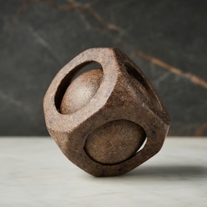 Puede incluir: Una escultura geométrica marrón con un marco en forma de cubo y dos elementos esféricos visibles a través de aberturas circulares. La escultura está hecha de un material texturizado similar a la piedra.