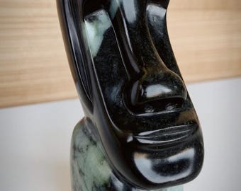Könnte beinhalten: Eine polierte, schwarze Skulptur eines Moai-Kopfes mit glatter, glänzender Oberfläche. Der Sockel hat ein gesprenkeltes grün-graues Muster. Die Skulptur ist eine Darstellung der ikonischen Osterinsel-Statuen.
