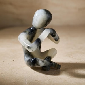 Könnte beinhalten: Kleine, sitzende Figur aus Stein, die eine menschenähnliche Form mit erhobenen Armen darstellt. Die Skulptur hat ein marmoriertes Aussehen mit schwarz-weißen Mustern. Die Figur befindet sich auf einer neutral getönten Oberfläche.