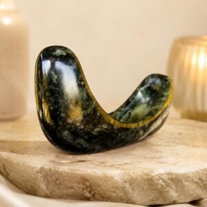 May include: A polished, crescent-shaped stone bowl featuring a deep forest green color with mottled black and gold veining. The smooth, curved object has a concave center and a glossy finish, showcasing natural mineral patterns throughout its surface.