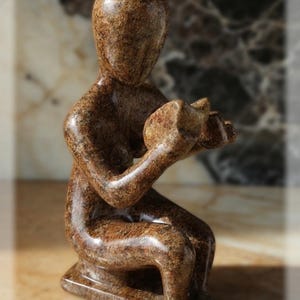 Könnte beinhalten: Eine braune Steinskulptur einer sitzenden Figur, die einen Gegenstand hält. Die Figur hat eine glatte, abgerundete Form mit einer strukturierten Oberfläche. Die Skulptur steht auf einem rechteckigen Sockel und hat ein poliertes Aussehen.