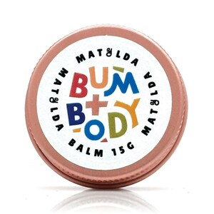 Op de afbeelding: Een ronde, roségouden blik van Matilda Bum + Body Balm. Het witte etiket toont de merknaam en productnaam in kleurrijke, speelse letters. De balsem weegt 15g.