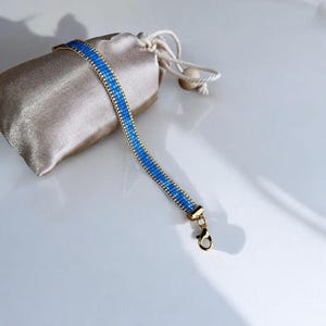 May include: A blue beaded bracelet with gold-colored accents and a clasp. The bracelet is next to a small, beige drawstring pouch. The bracelet's blue beads are arranged in a linear pattern, creating a vibrant contrast against the gold.