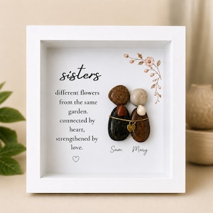 Peut inclure: Un cadre carré blanc contenant une œuvre d'art en galets représentant deux silhouettes formées de pierres lisses. Le texte indique : sisters different flowers from the same garden. connected by heart, strengthened by love. Les noms Sam et Mary sont inscrits.