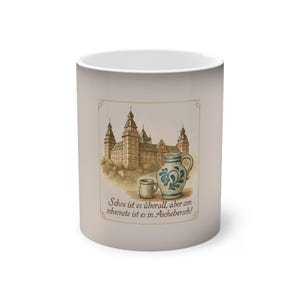 Puede incluir: Taza de cerámica beige con una ilustración de acuarela de un castillo, una taza pequeña y una jarra decorada en azul y blanco. La taza tiene texto en alemán. La taza es cilíndrica y tiene un interior blanco.