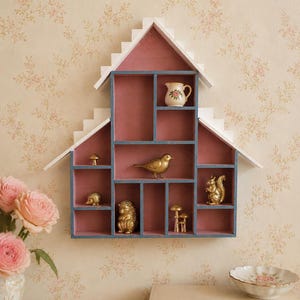 Pode incluir: Prateleira de parede em forma de casa, com interior rosa e moldura branca, contendo figuras douradas em miniatura de um pássaro, esquilo, ouriço e cogumelos. Uma pequena jarra com design floral também está presente. Montada em papel de parede floral.