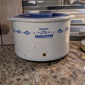 Puede incluir: Una olla de cocción lenta de cerámica Rival Crock-ette vintage. La olla de color crema tiene una tapa azul y está decorada con detalles florales azules. El logotipo de Crock-ette se muestra en la parte delantera. Una tabla de cortar de madera y una lámpara están en el fondo.