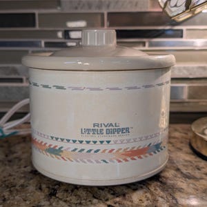 May include: A white ceramic mug with a black handle and the text "But First, Coffee" in a bold, black font. The mug is set against a blurred background, suggesting a cosy setting.