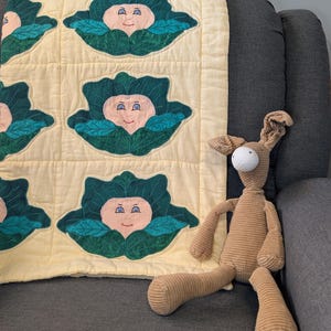 Pode incluir: Um edredom de bebê feito à mão com um padrão repetido de rostos sorridentes de repolho em um fundo amarelo. Um bicho de pelúcia marrom está ao lado do edredom em uma cadeira cinza.