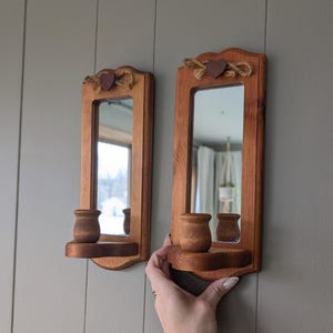 May include: Two wooden wall sconces with mirrors. Each sconce has a rectangular mirror, a small shelf, and a decorative heart and rope design at the top. The sconces are made of stained wood and are mounted on a gray wall.