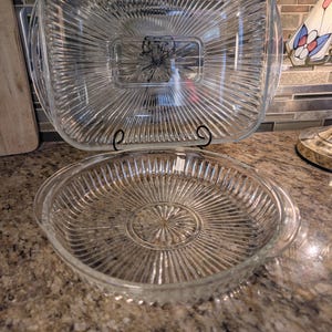 May include: Two clear glass serving dishes. The top dish is square with a rectangular centre and handles. The bottom dish is round with handles. Both dishes have a ribbed design radiating from the centre.