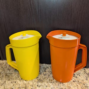 Op de afbeelding: Een felgele kan en een levendige oranje kan, beide voorzien van een wit deksel met een kleine centrale knop en een stevig handvat aan de zijkant. Beide containers hebben een gladde, effen afwerking en een brede schenktuit aan de bovenrand.