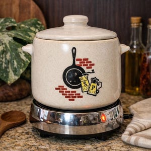 May include: A beige ceramic pot with a lid, featuring a black skillet and salt and pepper shakers graphic. The pot sits on a chrome electric burner with a red indicator light, ideal for cooking. The background includes a wooden cutting board and bottles.