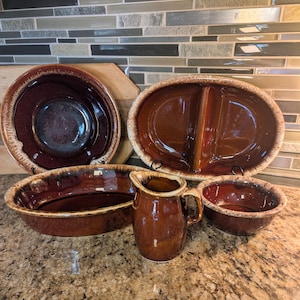 May include: A collection of brown ceramic serving dishes and a pitcher. The set includes a large round bowl, an oval divided platter, a long oval dish, a small pitcher, and a small bowl. The edges have a lighter brown trim.