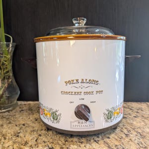 Può includere: Una pentola a cottura lenta bianca H&H Appliances Poke Along con coperchio in vetro trasparente, manici laterali marroni e una manopola di controllo marrone. Il fronte presenta il testo Poke Along Crockery Cook Pot e H&H Appliances con un bordo floreale.