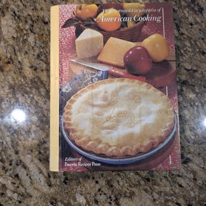 May include: A hardcover book titled The Illustrated Encyclopedia of American Cooking by Editors of Favorite Recipes Press. The cover features a photograph of a baked pie, various cheeses, a red apple, and a basket of fruit on a red patterned cloth.