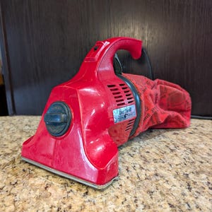 May include: A red handheld Dirt Devil vacuum cleaner featuring a black circular dial on the front, a top handle, and a red fabric dust bag with repeating black text patterns. The device has a wide intake nozzle and a black power cord extending out.