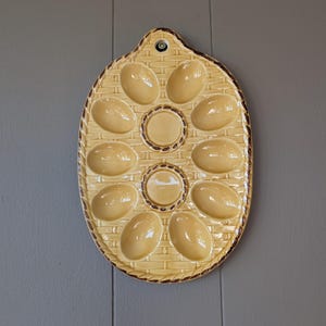 Peut inclure: Un porte-œufs ovale vintage en céramique. Le support jaune clair présente un motif de panier tressé et une bordure marron. Il comporte 10 empreintes en forme d'œuf et deux empreintes circulaires. Un petit trou est en haut pour accrocher.