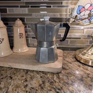 May include: A vintage silver stovetop espresso maker with a black handle and white knob. The coffee pot sits on a wooden chopping board, next to two beige ceramic spice jars. The background features a mosaic tile backsplash.