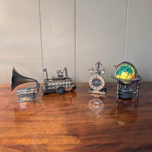 Puede incluir: Una colección de cuatro sacapuntas de metal en miniatura que incluye un gramófono, un barco de vapor, un reloj con una campana en la parte superior y un globo terráqueo sobre un soporte. Cada objeto presenta detalles intrincados y acabado metálico.