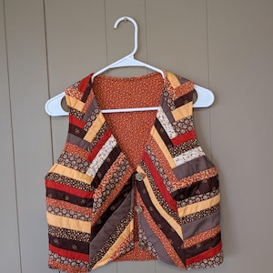 May include: A sleeveless vest featuring a quilted herringbone pattern in shades of brown, orange, and yellow. The garment has a V-neckline, three circular buttons down the front, and a contrasting orange floral print lining visible on the interior.